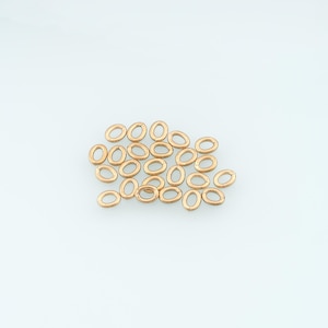 May include: A collection of small, oval-shaped jump rings with a metallic gold finish. These jewelry components are arranged in a loose cluster, each featuring a thin, circular wire construction with a visible opening for connecting jewelry pieces.