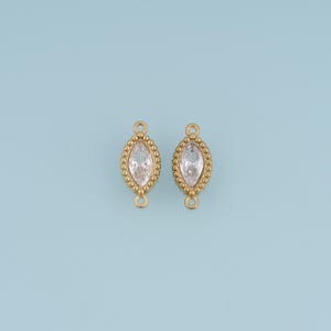 May include: Two gold-toned jewellery connectors featuring a central marquise-cut clear gemstone surrounded by a beaded metal border. Each connector has a small circular loop at the top and bottom for attachment, showcasing a symmetrical design.