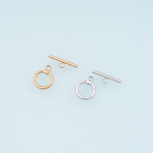 May include: Two toggle clasps, one gold-toned and one silver-toned, each with a circular ring and a straight bar. The clasps are displayed on a light blue background, showcasing their design and finish. Ideal for jewelry making.
