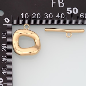 May include: Gold-colored jewelry findings, including a large, irregular-shaped pendant with a loop and a small bar with a loop. The pendant has a smooth, polished surface. The bar is straight with a loop at one end.