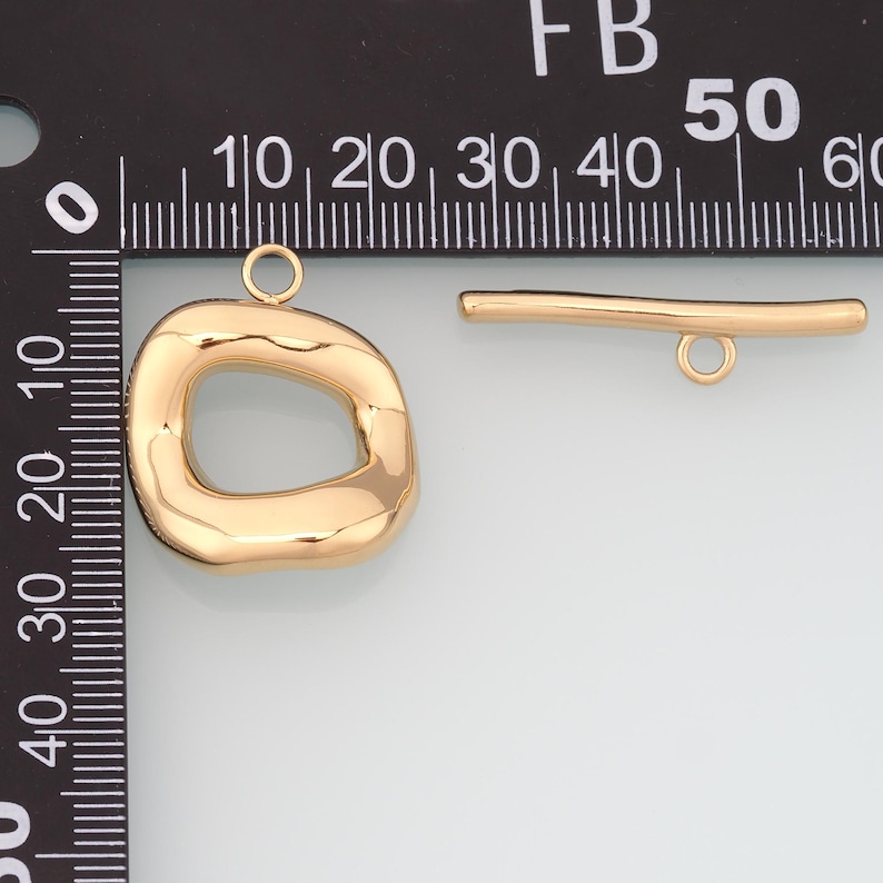 May include: Gold-colored jewelry findings, including a large, irregular-shaped pendant with a loop and a small bar with a loop. The pendant has a smooth, polished surface. The bar is straight with a loop at one end.