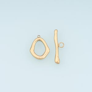 May include: Gold-toned toggle clasp jewelry findings. The clasp features an irregular oval loop and a curved bar with a small loop. The jewelry findings are displayed against a light blue background.