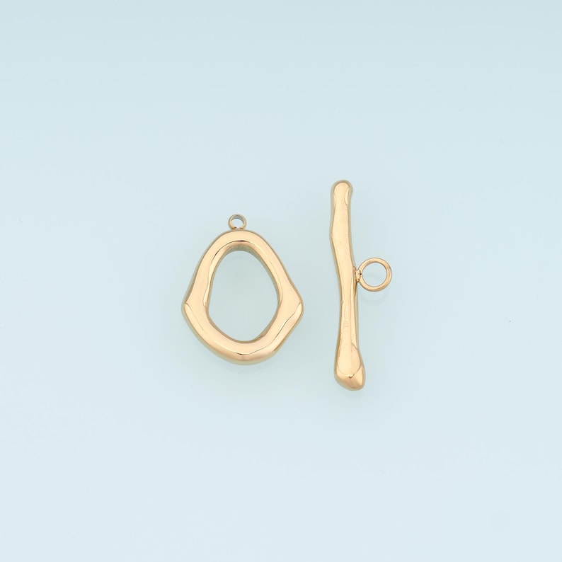 May include: Gold-toned toggle clasp jewelry findings. The clasp features an irregular oval loop and a curved bar with a small loop. The jewelry findings are displayed against a light blue background.