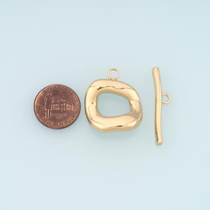 May include: Gold-toned toggle clasp jewelry findings. The clasp features an irregular, rounded rectangle and a straight bar with a loop. A US one-cent coin is included for size reference.