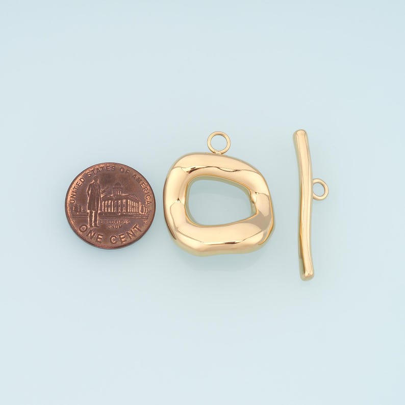 May include: Gold-toned toggle clasp jewelry findings. The clasp features an irregular, rounded rectangle and a straight bar with a loop. A US one-cent coin is included for size reference.