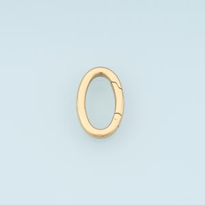 May include: A gold-colored oval-shaped clasp, open on one side, against a light blue background. The clasp has a smooth, polished surface and a hinged mechanism for opening and closing. The design is simple and elegant.