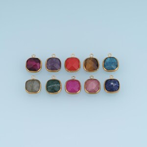 Puede incluir: Un conjunto de diez dijes cuadrados para joyería, cada uno con una piedra preciosa facetada engastada en un marco de metal dorado con un pequeño lazo circular en la parte superior. Las piedras muestran una variedad de colores que incluyen rojo, morado, azul, verde y gris.