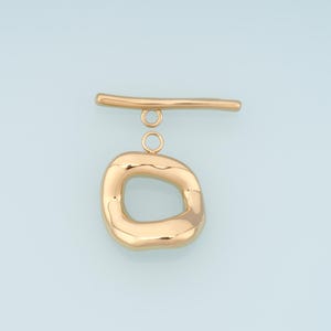 May include: A gold-colored toggle clasp with a unique, organic shape. The clasp features a rounded, irregular oval with a bar and two small connecting rings. The metal has a polished, reflective surface, set against a light blue background.
