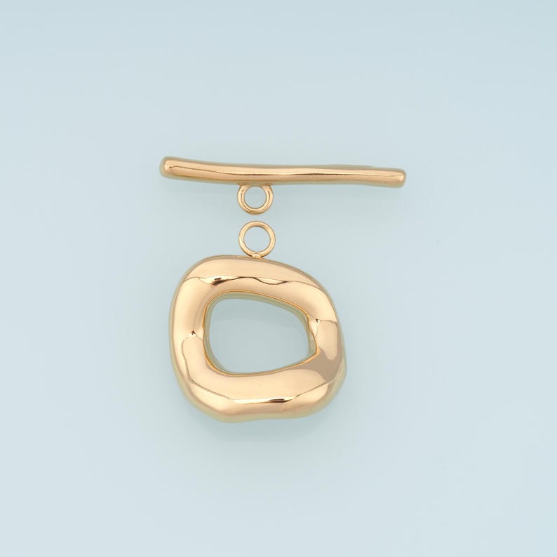 May include: A gold-colored toggle clasp with a unique, organic shape. The clasp features a rounded, irregular oval with a bar and two small connecting rings. The metal has a polished, reflective surface, set against a light blue background.