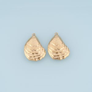 May include: Two gold-toned metal charms shaped like oyster shells. Each charm features a textured, rippled surface with a small circular loop at the top for attachment. The pieces have a metallic finish and a distinct, organic shell-like form.