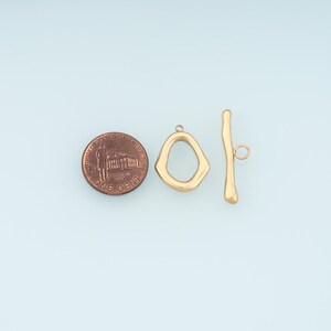 May include: Gold-colored toggle clasp jewelry findings. The clasp includes an irregular-shaped ring and a bar with a small loop. A US penny is included for size reference.