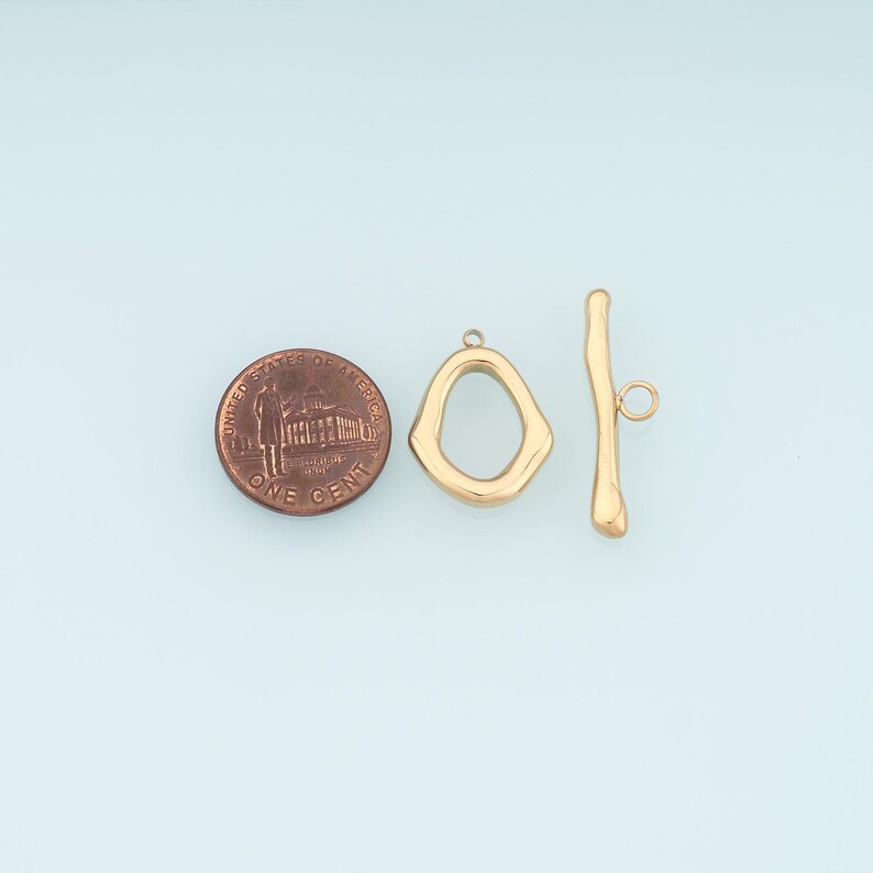May include: Gold-colored toggle clasp jewelry findings. The clasp includes an irregular-shaped ring and a bar with a small loop. A US penny is included for size reference.