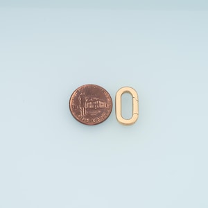 Może przedstawiać: Złota owalna klamra obok amerykańskiego centa. Klamra jest otwarta, pokazując wnętrze. Cent ma kolor miedziany i przedstawia profil Abrahama Lincolna. Tło jest jasnoniebieskie.