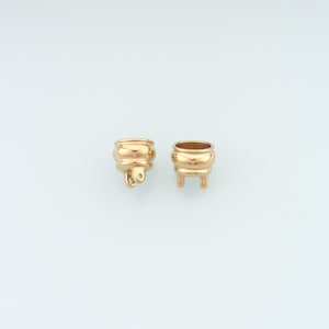 Pode incluir: Duas pequenas peças de joalharia em metal dourado, especificamente tampas de contas ou terminais de cordão. Cada peça apresenta uma superfície exterior arredondada e lisa com um centro oco e um pequeno laço ou ponto de fixação na base.