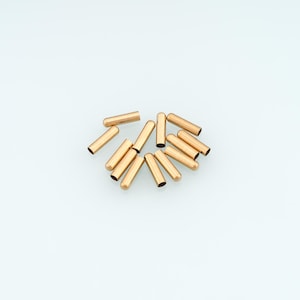 May include: A collection of twelve small, cylindrical, gold-toned metal crimp covers. Each piece features a smooth, polished surface with a hollow interior and a rounded, closed end, designed for jewellery making and finishing cord ends.