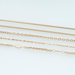 May include: Five distinct gold-toned metal chains arranged horizontally. The collection includes a square box chain, a braided wheat chain, a textured oval link chain, a delicate bar link chain, and a mixed link chain with elongated ovals.