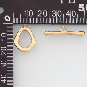 May include: Two gold-colored toggle clasps. One is an irregular oval shape with a loop at the top. The other is a bar with a loop. Both clasps are shiny and appear to be made of metal. The clasps are shown next to a ruler.