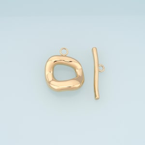 May include: Gold-colored toggle clasp jewelry findings. The clasp features a rounded, irregular oval shape with a loop, and a straight bar with a loop. The jewelry findings are on a light blue background.