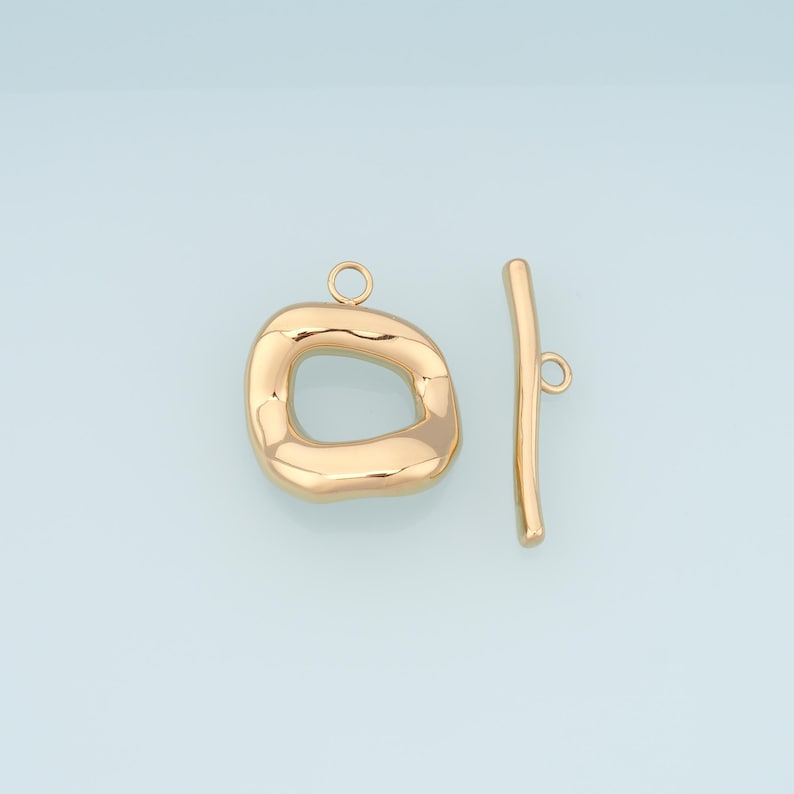 May include: Gold-colored toggle clasp jewelry findings. The clasp features a rounded, irregular oval shape with a loop, and a straight bar with a loop. The jewelry findings are on a light blue background.