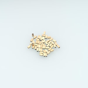 May include: A collection of small, rectangular, gold-colored metal beads with a smooth, polished finish. Each bead features a central hole running through its length, arranged in a loose, scattered cluster on a plain, light-colored background.