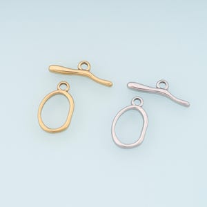May include: Two sets of jewelry clasps, one gold-toned and one silver-toned, against a light blue background. Each set includes an oval-shaped ring and a curved bar with a loop for attachment. Ideal for jewelry making.