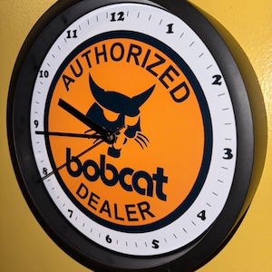 Puede incluir: Reloj de pared redondo negro con esfera blanca y manecillas negras. El reloj presenta las palabras "AUTHORIZED bobcat DEALER" en un diseño circular con fondo naranja y un logotipo negro de bobcat.