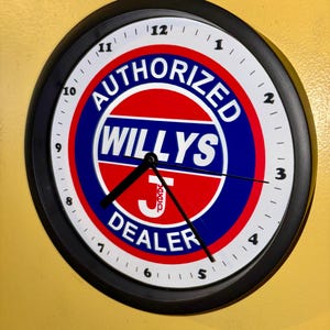 May include: A round wall clock with a black frame and a white face featuring black numerals. The centre displays a red and blue circular logo with the text AUTHORIZED WILLYS DEALER and a white J logo. Three black clock hands indicate the time.