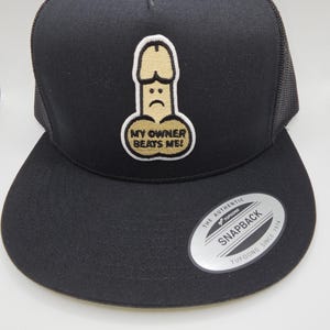 May include: Black trucker cap featuring a beige and gold embroidered design of a sad penis with the text "MY OWNER BEATS ME!". The cap has a curved peak and a mesh back.