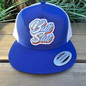 May include: A blue and white trucker hat with the text "Butt Slut" in a colorful, embroidered design. The hat features a curved brim and a snapback closure. The hat has a "Snapback" tag.