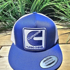 Peut inclure: Casquette de camionneur bleue et blanche avec un écusson du logo Cummins. La casquette a une visière incurvée, un dos en filet blanc et une fermeture à pression. L'écusson comprend les mots "Cummins" et "Turbo Diesel".