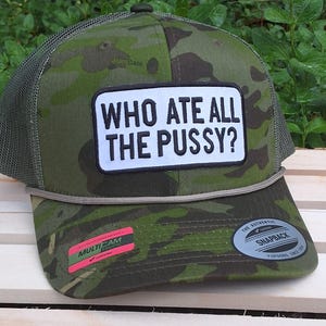 May include: A camouflage trucker hat with a white patch that reads "WHO ATE ALL THE PUSSY?". The hat has a green mesh back and a curved brim. The hat has a snapback closure and a rope detail.