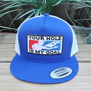 Op de afbeelding: Blauwe en witte truckerpet met een blauwe klep en een witte mesh achterkant. De pet heeft een patch met de tekst "YOUR HOLE IS MY GOAL" en een afbeelding van een persoon die een spel speelt. Met snapback sluiting.