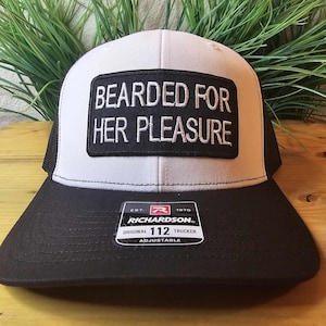 May include: A black and white trucker hat with the text "BEARDED FOR HER PLEASURE" on a black patch. The hat has a black brim and mesh back, with a Richardson 112 tag.