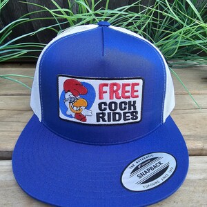 Puede incluir: Gorra de camionero azul y blanca con visera plana. La gorra presenta un parche con un gallo de dibujos animados y las palabras "FREE COCK RIDES". La gorra tiene un cierre a presión y una etiqueta Yupoong.