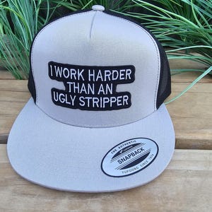 Puede incluir: Gorra de camionero gris y negra con el texto "I WORK HARDER THAN AN UGLY STRIPPER" en un parche negro. La gorra tiene una visera curva y un cierre snapback. La marca "Yupoong" es visible.