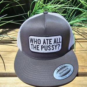 May include: A grey and white trucker cap with a flat peak. The cap has a rectangular patch with the text "WHO ATE ALL THE PUSSY?" in white. It features a snapback closure and a Yupoong label.