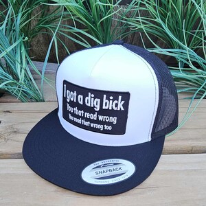 May include: A black and white trucker cap with a white front panel and a black brim and mesh back. The front panel has a black rectangle with the text "I got a dig bick You that read wrong You read that wrong too."