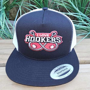 Può includere: Cappellino da camionista nero e bianco con la scritta "I Love Hookers" in un design rosso e bianco. Il cappellino ha una visiera curva e una chiusura a scatto. È un Yupoong Snapback.