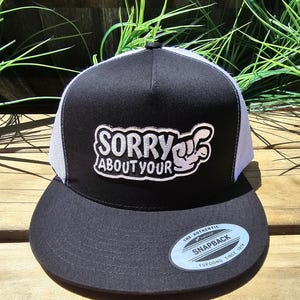 Puede incluir: Gorra snapback negra y blanca con el texto "SORRY ABOUT YOUR" en blanco. La gorra tiene una visera y una corona negras, con una parte trasera de malla blanca. Una pequeña etiqueta ovalada en la visera dice "SNAPBACK".