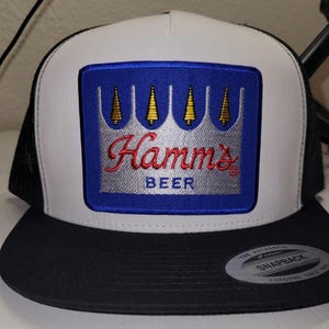 May include: A black, white, and blue trucker hat with the Hamm's Beer logo. The logo is a rectangular patch with a blue background, a silver crown, and the word "Hamm's" in red script. The hat has a snapback closure.