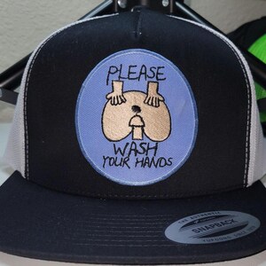 May include: Black and white trucker hat with a blue patch featuring a humorous illustration and the text "PLEASE WASH YOUR HANDS". The hat has a curved peak and a snapback closure.
