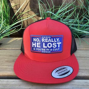 May include: A red and black Snapback hat with the text "NO, REALLY. HE LOST & YOU'RE IN A CULT" on a blue patch. The hat has a flat brim and a mesh back. The brand name "YUPOONG" is visible on the brim.