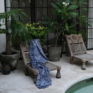 May include: Two low-profile wooden chairs with carved backrests and turned legs are positioned near a swimming pool. A blue fabric with a white geometric pattern is draped over one chair. Several potted green plants surround the seating area.