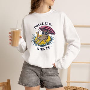 May include: A white long-sleeve sweatshirt featuring a graphic of a brown cat wearing sunglasses, relaxing on a yellow pool float under a pink and black striped umbrella, holding a drink. The text DOLCE FAR NIENTE is printed in black above and below.