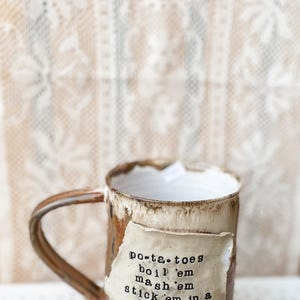 Op de afbeelding: Een handgemaakte keramische mok met een bruin en witte glazuur. Een gescheurd papieren label is aan de mok bevestigd met de tekst "po-ta-toes boil 'em mash 'em stick 'em in a stew". De mok heeft een gebogen handvat.