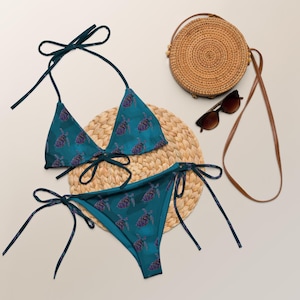 Puede incluir: Conjunto de bikini turquesa con estampado de tortugas, con top triangular y braguita con lazos. En la imagen también hay un bolso redondo tejido, gafas de sol y una esterilla tejida, creando una estética playera.