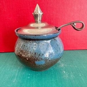 May include: A ceramic sugar bowl with a blue and brown speckled glaze. It has a silver-colored lid and a matching spoon. The lid is topped with a decorative finial. The bowl is round and sits on a green surface.