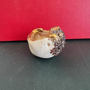 May include: A small, handcrafted ceramic bowl with a textured, off-white exterior. The rim and a portion of the exterior are accented with gold. The bowl's right side features a textured, dark brown and black design. The bowl is on a dark green surface.