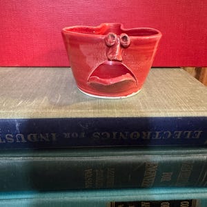 May include: A handmade, red ceramic bowl with a face design. The bowl has a wide, open mouth and two eye-like features above. The bowl is sitting on top of a stack of old books with a red background.