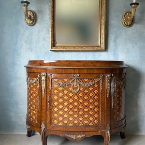 May include: A vintage wooden cabinet with a curved front, featuring intricate inlaid patterns and ornate brass accents. A gold-framed mirror hangs above, flanked by two gold sconces with white shades. The cabinet has a height of approximately 36 inches.